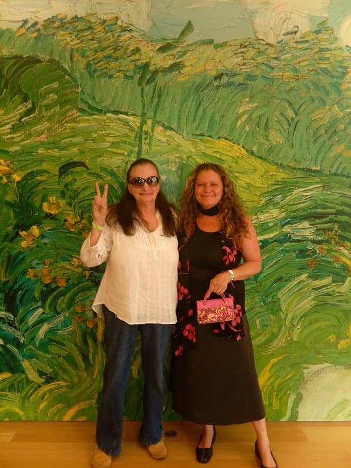 Nancy Sinkora smiling and holding up a peace sign in front of green artwork